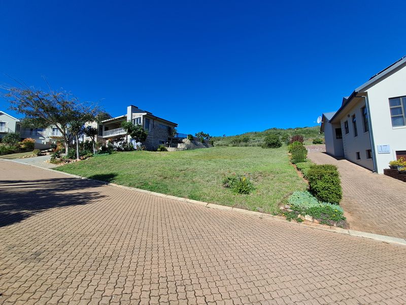 A vacant residential plot situated between established homes, featuring a gentle slope, manicured lawn, and paved driveway access.