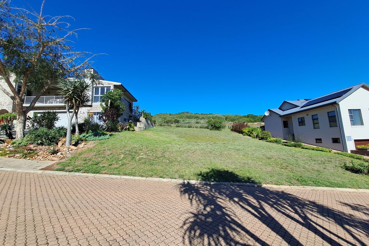 A vacant residential plot situated between two existing homes, featuring a gentle slope and paved street frontage in a developed neighborhood.
