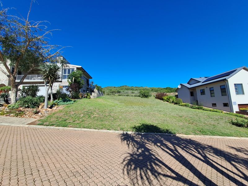 A vacant residential plot situated between two existing homes, featuring a gentle slope and paved street frontage in a developed neighborhood.