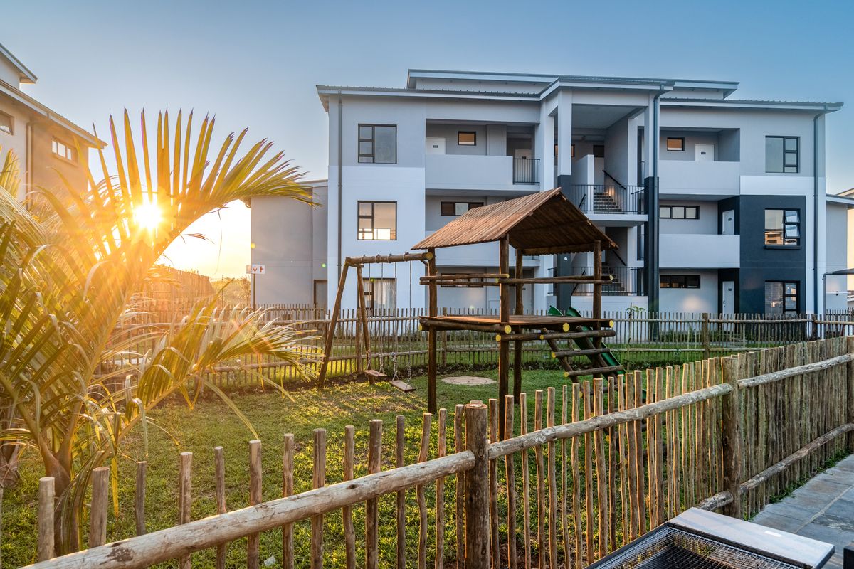 A well-maintained apartment complex exterior featuring a landscaped garden area with a wooden play structure and secure picket fencing at sunset.