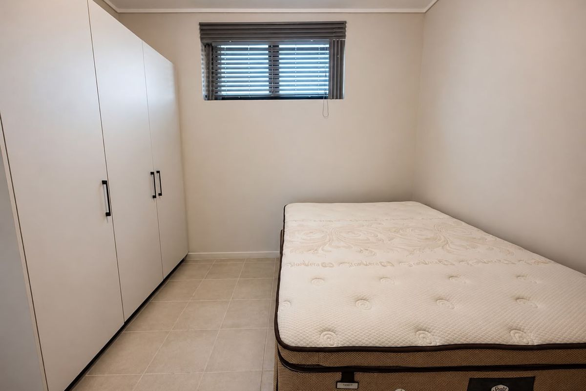A clean bedroom featuring neutral tiled flooring, a built-in wardrobe with modern handles, and a window fitted with venetian blinds.