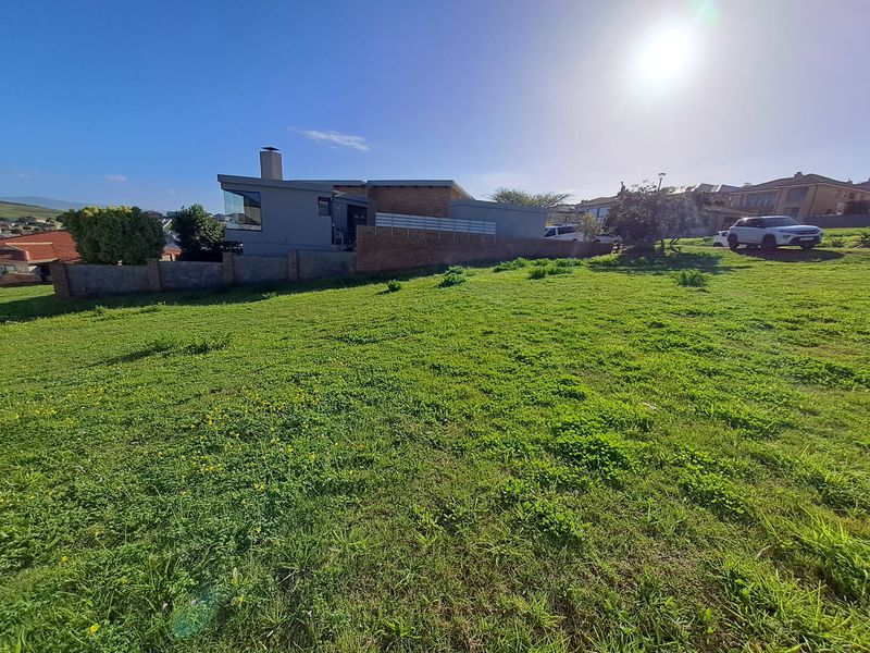 A view of a vacant residential plot featuring a grass lawn and a brick boundary wall, with neighboring properties visible in the background.
