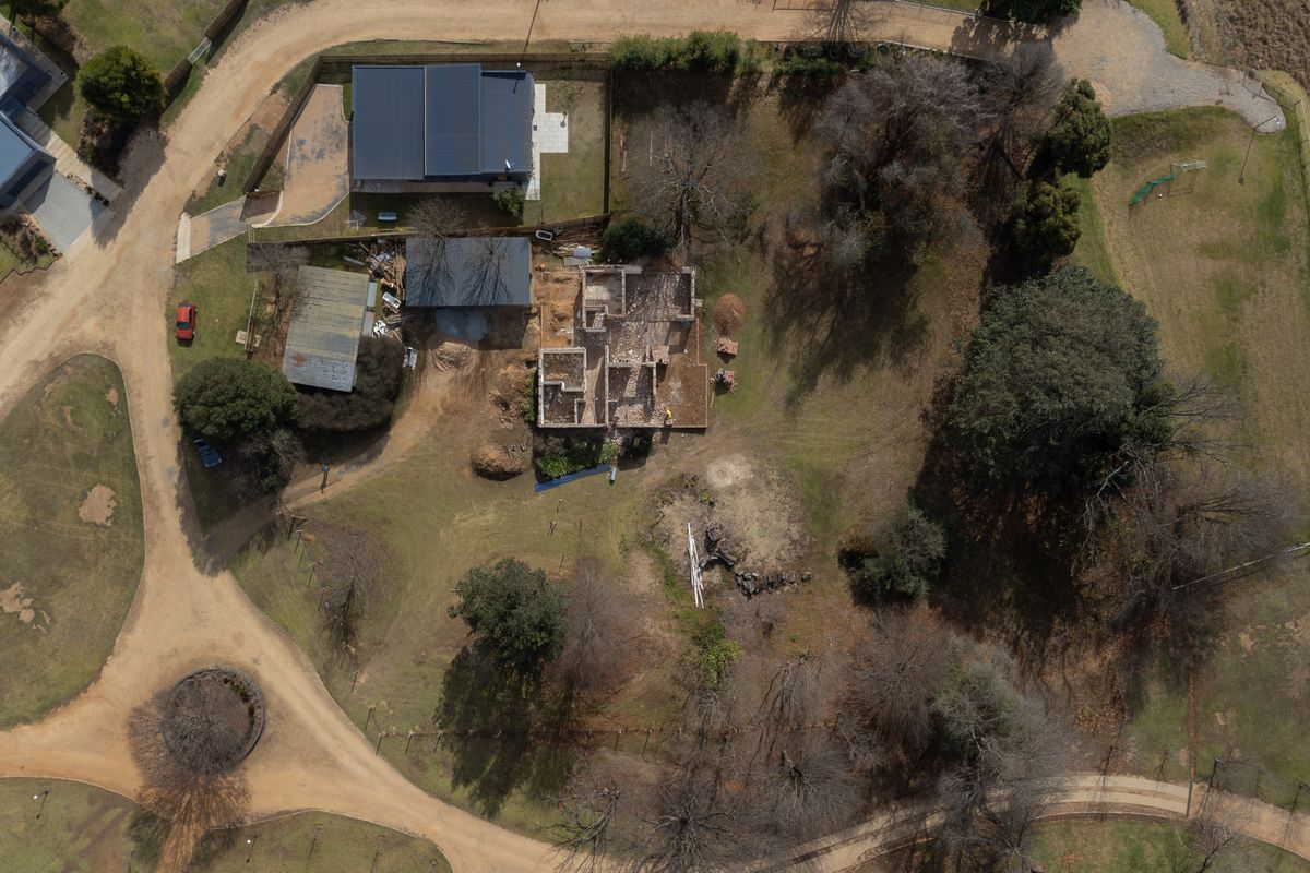 Aerial view of a residential plot featuring an existing outbuilding and a partially constructed building structure amidst mature trees and dirt access roads.