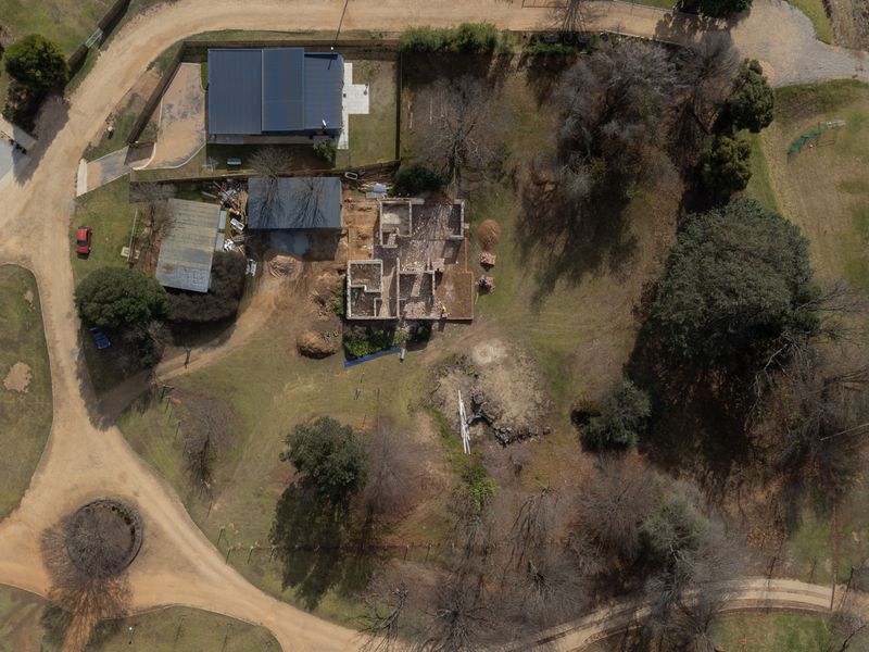 Aerial view of a residential plot featuring an existing outbuilding and a partially constructed building structure amidst mature trees and dirt access roads.