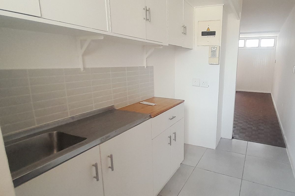 A functional kitchen area featuring white built-in cabinetry, a stainless steel sink, tiled backsplash, and neutral floor tiles, leading into an adjacent room.
