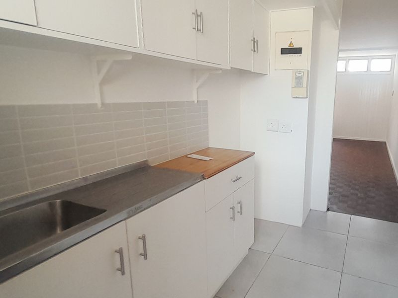 A functional kitchen area featuring white built-in cabinetry, a stainless steel sink, tiled backsplash, and neutral floor tiles, leading into an adjacent room.