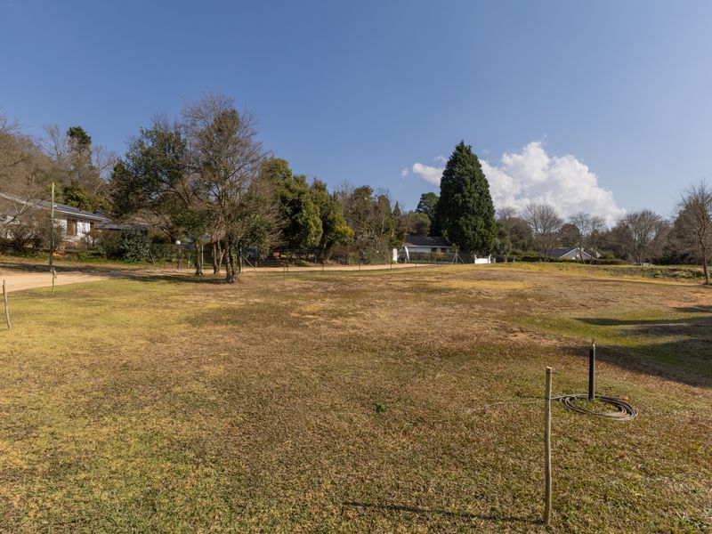 A spacious, open residential plot featuring established trees, a grass lawn, and perimeter fencing, offering a clear view of the surrounding neighborhood.