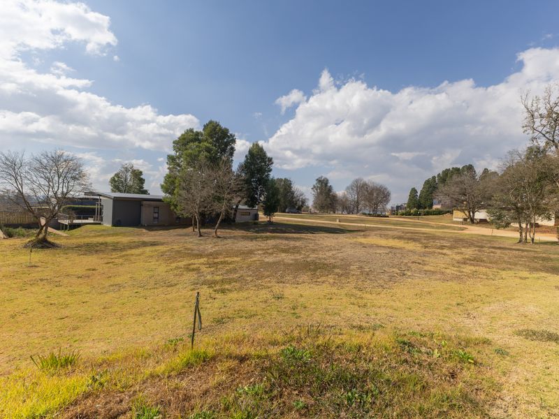 A wide-angle view of a spacious, open residential plot featuring natural grass cover and mature trees under a clear sky.