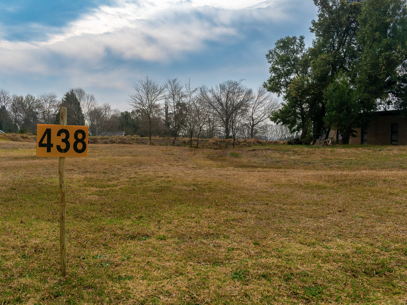 A vacant residential plot of land featuring open grass and mature trees, marked with a property number sign.