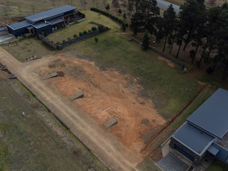 Aerial view of a vacant residential plot featuring cleared land, established retaining walls, and gravel driveway access situated between existing modern homes.