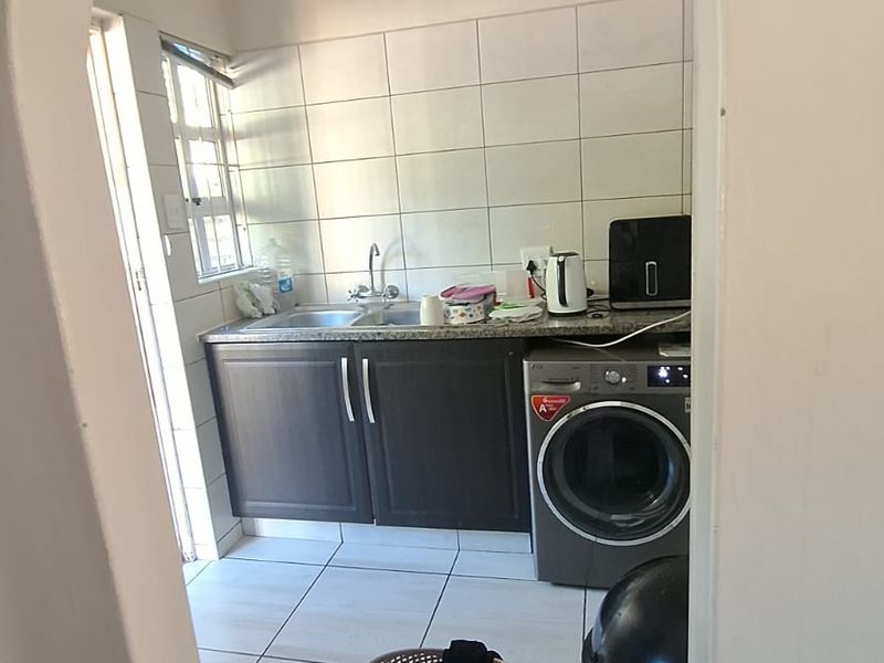A functional laundry area featuring built-in dark cabinetry, a stainless steel sink, tiled splashback, and tiled flooring, accessed through an arched doorway.