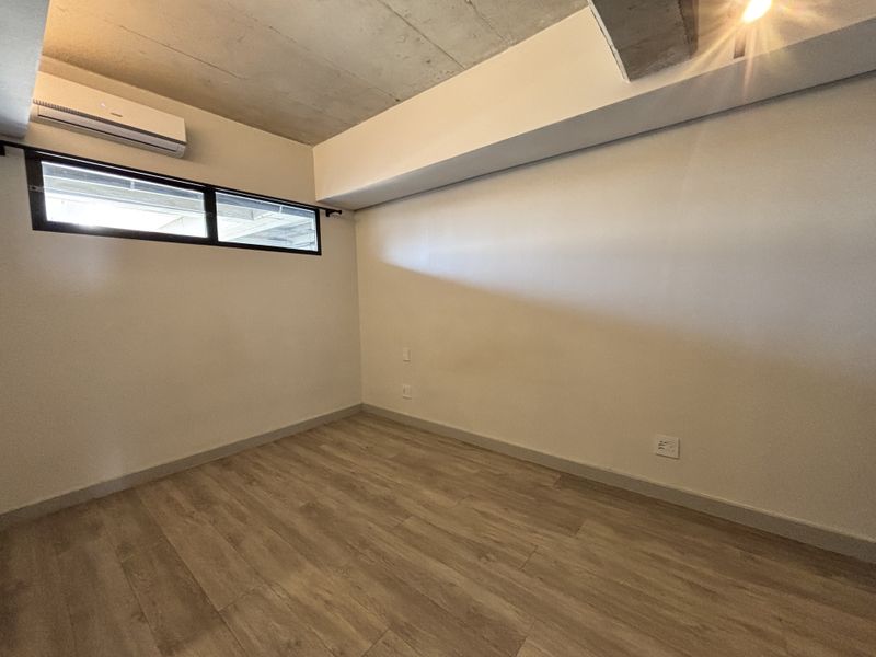 An empty bedroom featuring modern laminate flooring, an exposed concrete ceiling, and a high-level clerestory window with an integrated air conditioning unit.