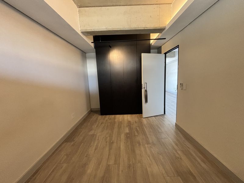 A modern, empty bedroom featuring wood-look tile flooring, a sleek dark built-in wardrobe, and an exposed concrete ceiling beam.