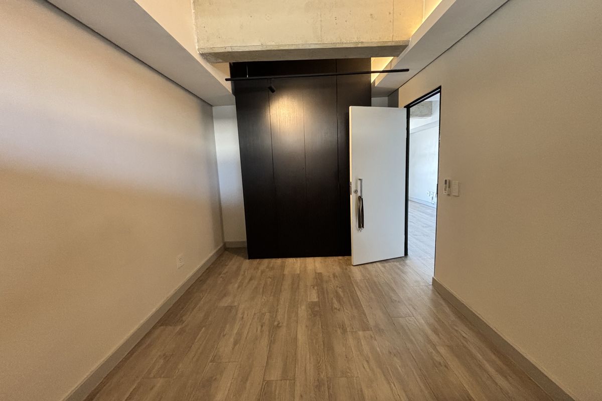 A modern, empty bedroom featuring wood-look tile flooring, a sleek dark built-in wardrobe, and an exposed concrete ceiling beam.