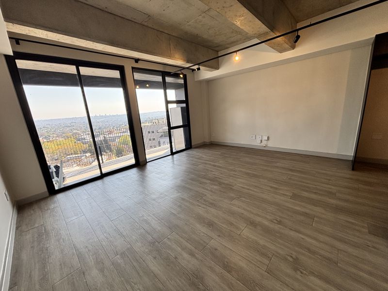 Spacious open-plan living area featuring modern wood-look tile flooring, exposed concrete ceilings, track lighting, and large sliding doors leading to a balcony with city views.