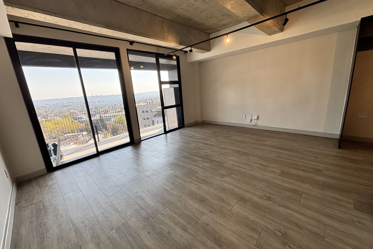 Spacious open-plan living area featuring modern wood-look tile flooring, exposed concrete ceilings, track lighting, and large sliding doors leading to a balcony with city views.