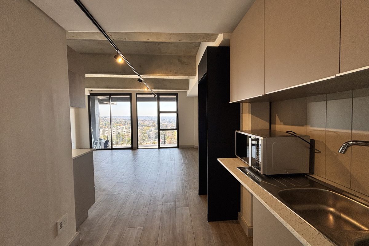 Modern open-plan apartment featuring an exposed concrete ceiling, sleek cabinetry, and large sliding doors leading to a balcony with expansive city views.