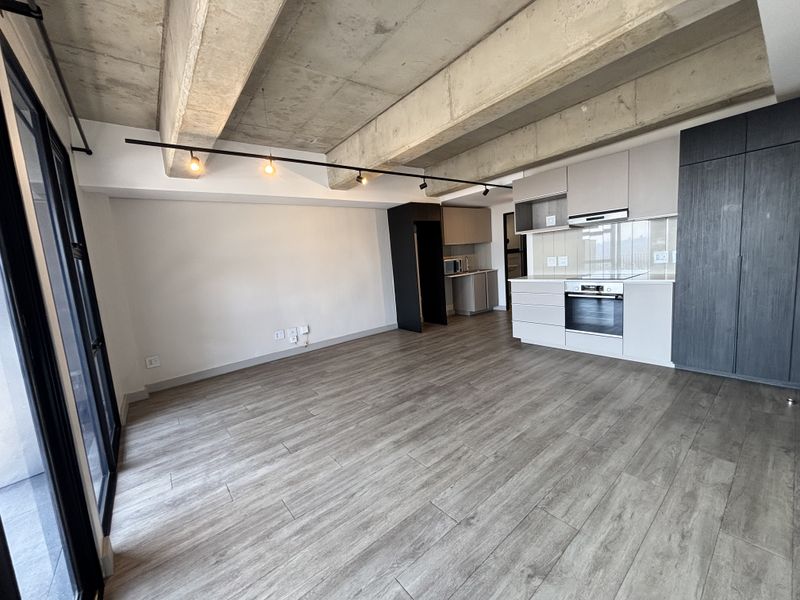 Modern open-plan living area featuring an exposed concrete ceiling, sleek built-in kitchen cabinetry with integrated appliances, and durable laminate flooring.