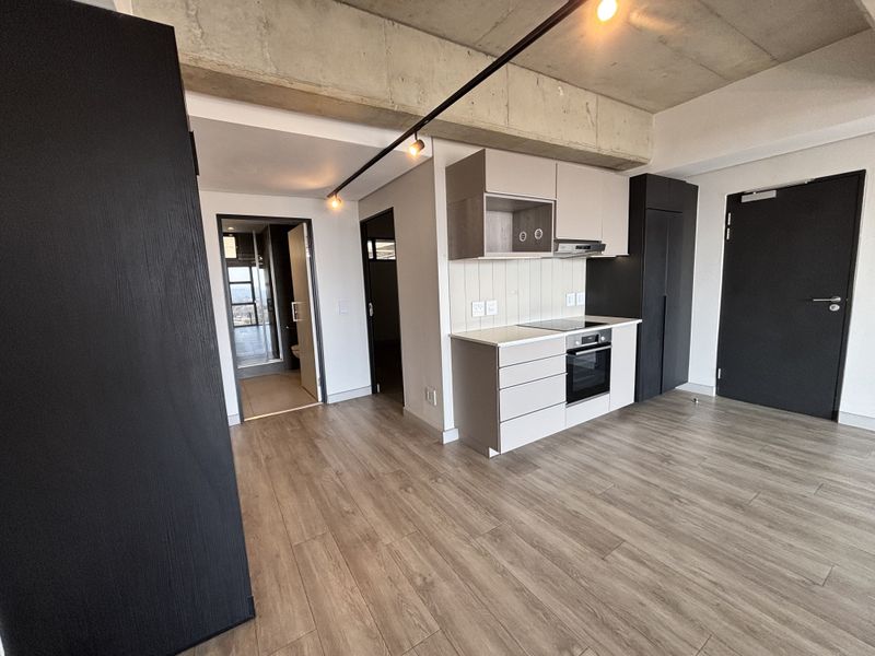 Modern open-plan apartment featuring an exposed concrete ceiling, sleek built-in kitchen cabinetry with integrated appliances, and durable laminate flooring throughout.