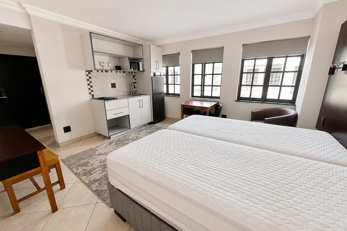 Open-plan studio apartment featuring a compact kitchenette with built-in cabinetry, tiled flooring, and large multi-pane windows providing ample natural light.