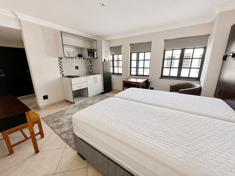 Open-plan studio apartment featuring a compact kitchenette with built-in cabinetry, tiled flooring, and large multi-pane windows providing ample natural light.