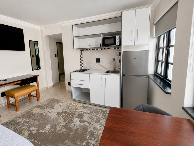 Compact open-plan kitchenette featuring modern white cabinetry, stone-look backsplash, built-in microwave, and hob, integrated into a functional living space with tiled flooring.