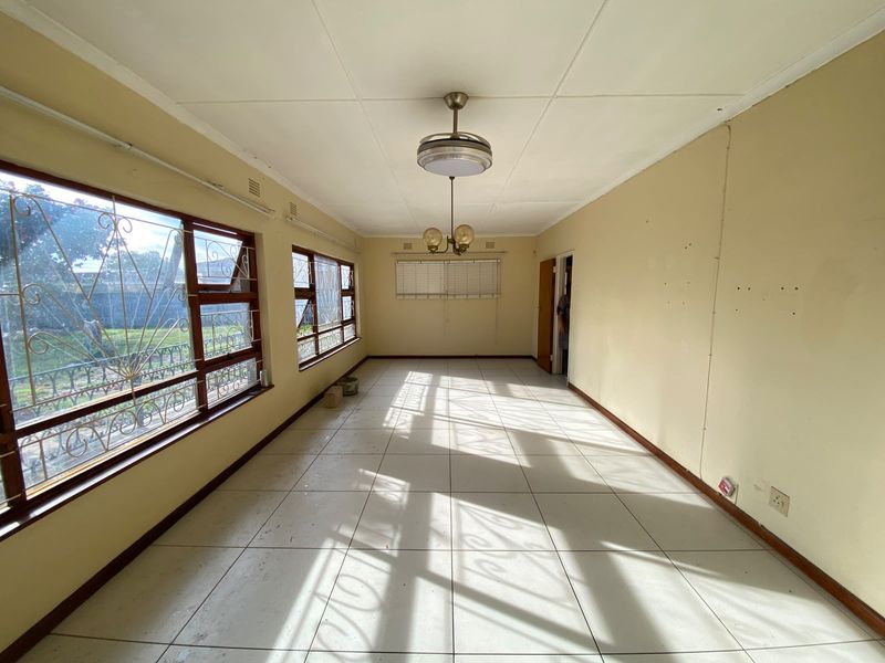 An empty, sunlit living area featuring tiled flooring, large windows with security bars, and dual ceiling light fixtures, ready for renovation or staging.