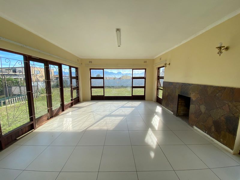 Spacious, light-filled living room featuring large windows with garden views, tiled flooring, and a stone-clad fireplace.