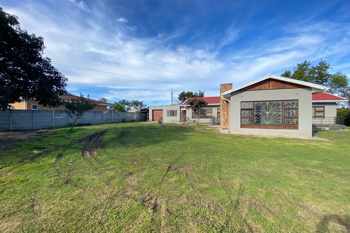 Rear view of the property featuring a large lawn, precast concrete boundary wall, and a house with a stone-clad chimney and metal roofing.