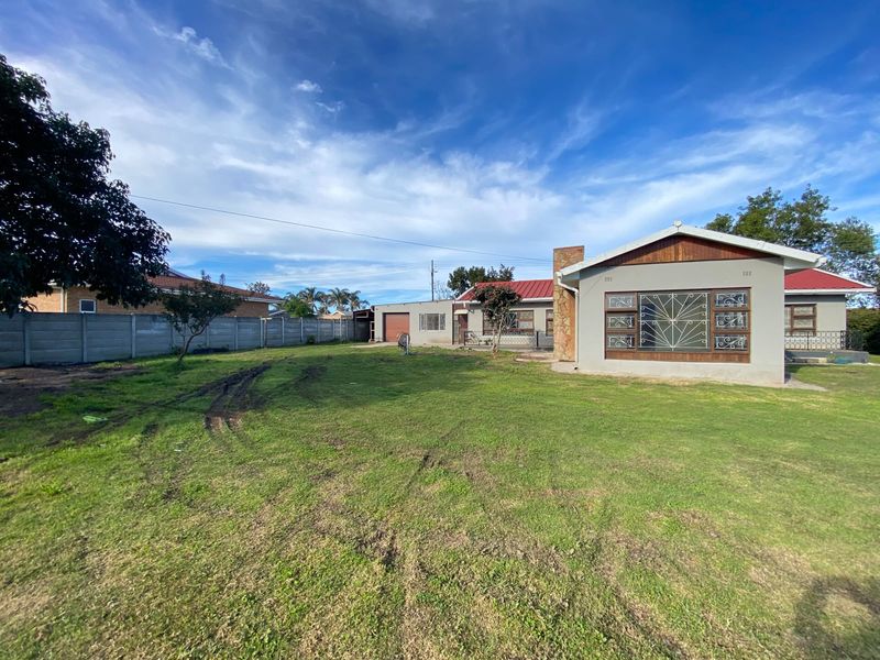 Rear view of the property featuring a large lawn, precast concrete boundary wall, and a house with a stone-clad chimney and metal roofing.