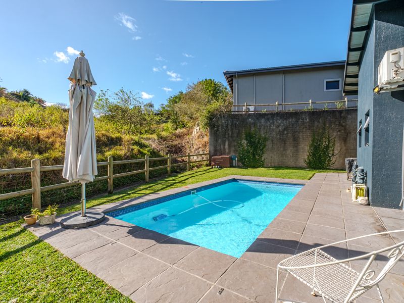 Outdoor pool area featuring a tiled surround, lawn space, and a retaining wall, set against a natural hillside backdrop.