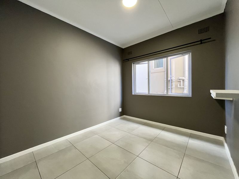 An empty bedroom featuring neutral grey walls, large format floor tiles, and a window with a curtain rod.