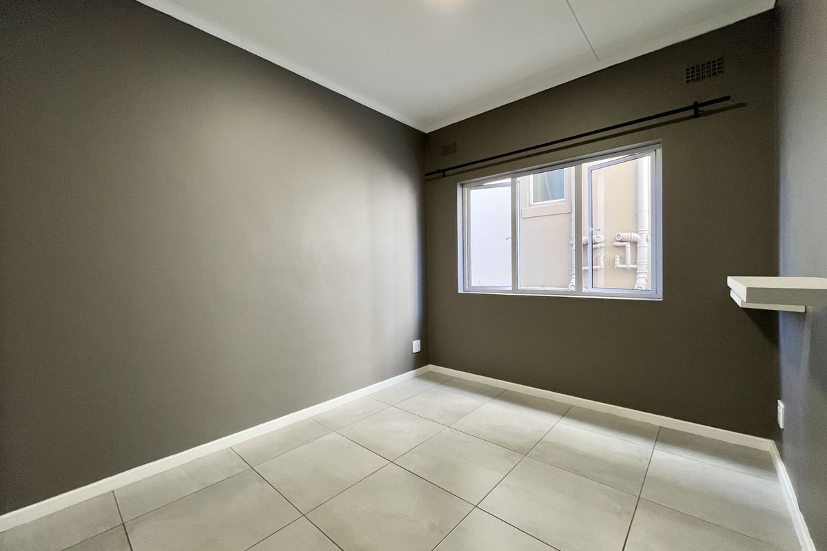An empty bedroom featuring neutral grey walls, large format floor tiles, and a window with a curtain rod.