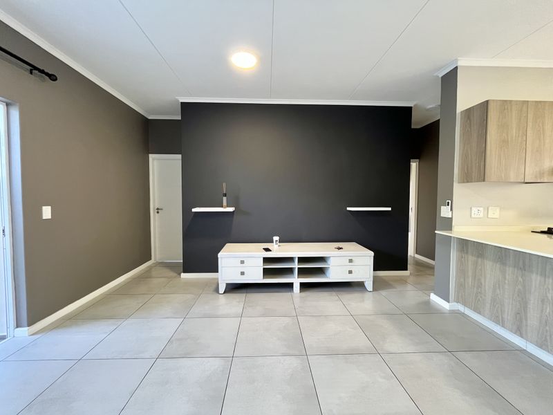 An open-plan living area featuring large format floor tiles, a modern kitchen with wood-finish cabinetry, and a dark accent wall with floating shelves.