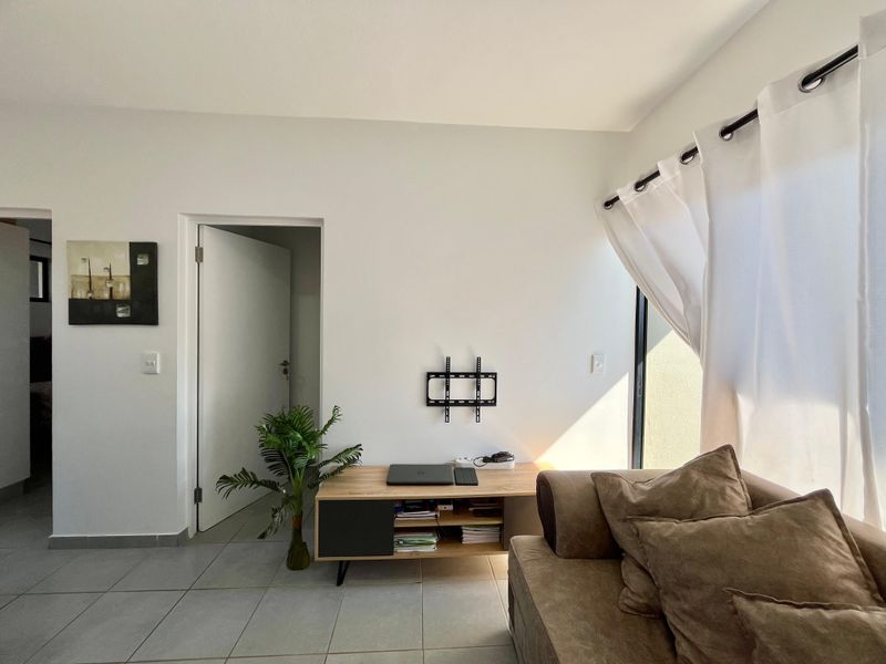 A bright living room featuring neutral tiled flooring, a wall-mounted TV bracket, and access to an outdoor area through a sliding glass door.