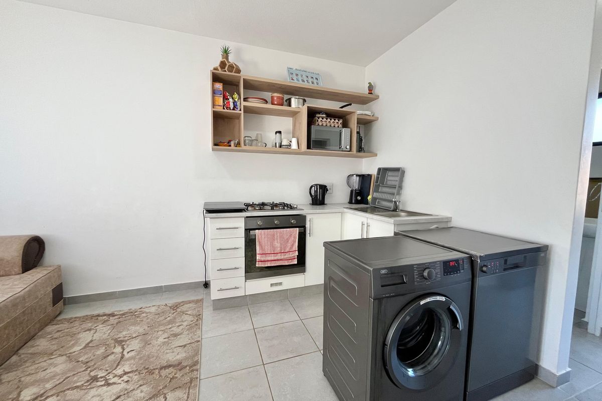 Compact kitchen area featuring built-in oven, gas hob, stainless steel sink, and open shelving, with tiled flooring and space for laundry appliances.