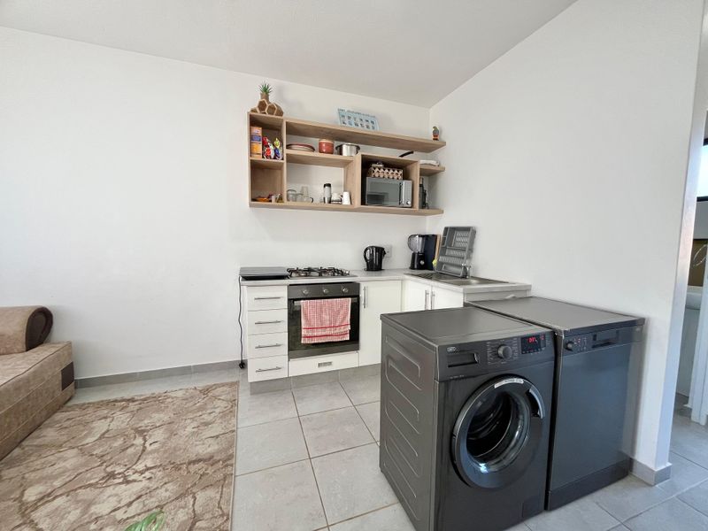 Compact kitchen area featuring built-in oven, gas hob, stainless steel sink, and open shelving, with tiled flooring and space for laundry appliances.
