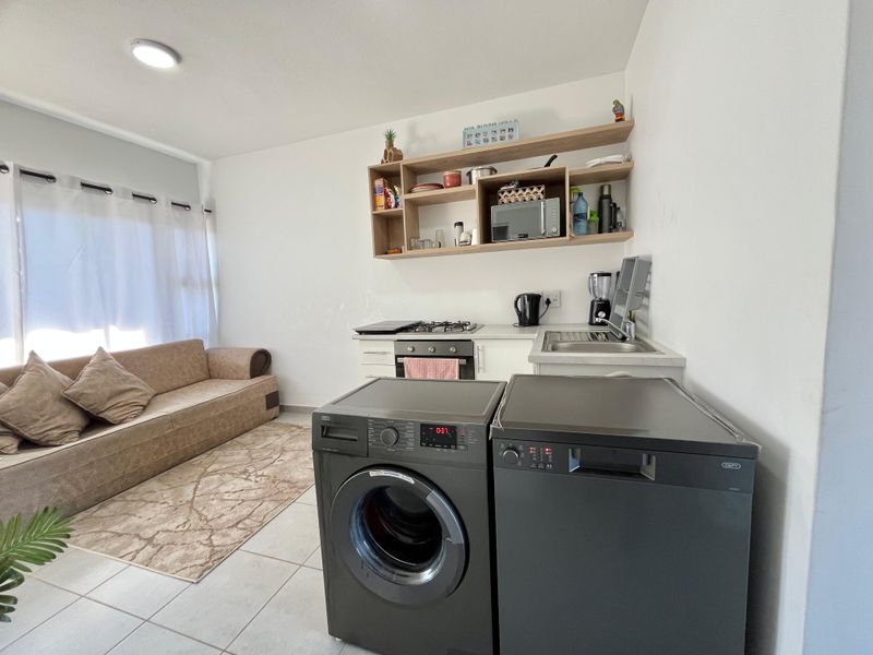 Compact open-plan living area featuring a kitchenette with built-in oven, gas hob, stainless steel sink, and open shelving, finished with tiled flooring.