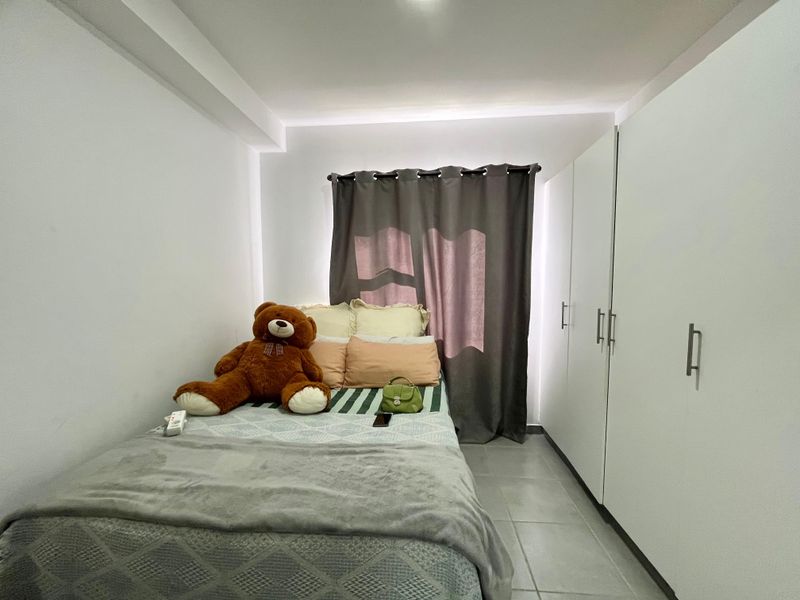 A compact bedroom featuring neutral tiled flooring, built-in white wardrobes, and recessed ceiling lighting, providing a clean and functional living space.