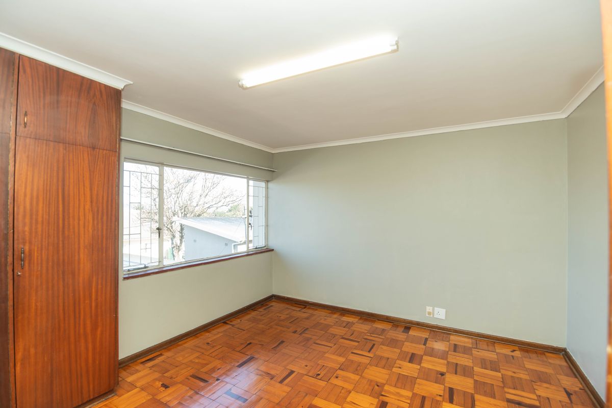 A bright, empty bedroom featuring classic parquet flooring, a built-in wooden wardrobe, and large windows with security bars.