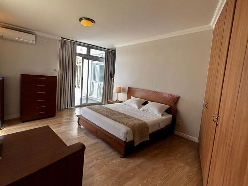 A well-maintained bedroom featuring laminate flooring, built-in wooden cupboards, a split-system air conditioner, and access to a private balcony through sliding glass doors.
