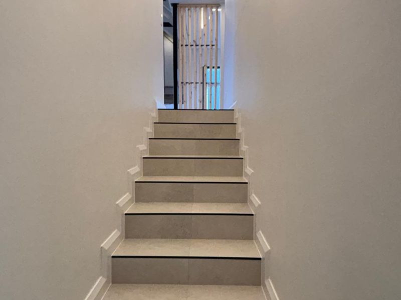 A modern, minimalist staircase featuring neutral tiled steps, clean white walls, and integrated linear LED lighting leading to a timber slat feature screen.