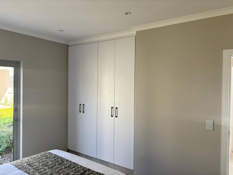 A well-maintained bedroom featuring neutral wall tones, built-in floor-to-ceiling cupboards, recessed lighting, and direct access to the garden via a sliding glass door.