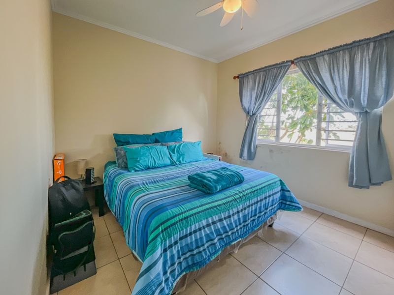 A bedroom featuring tiled flooring, a ceiling fan, and a window with natural light. Visible damp damage is present on the wall below the window.