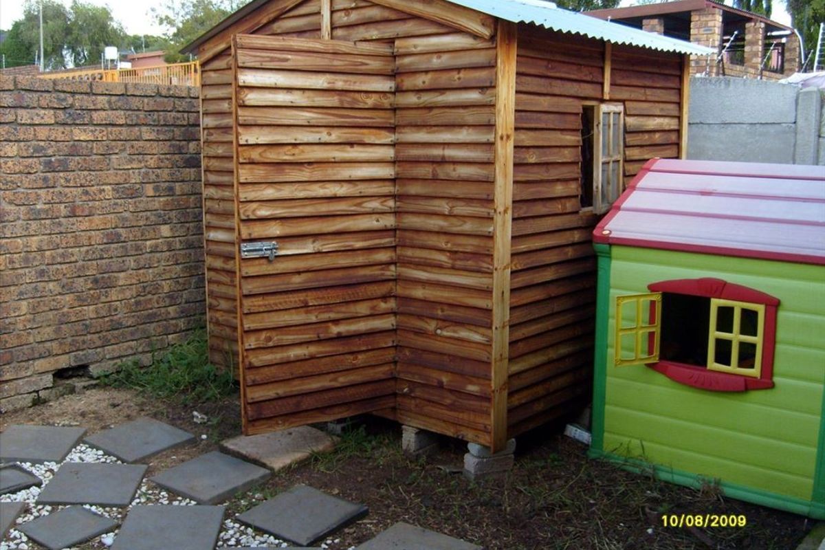 A wooden garden storage shed situated in the backyard, featuring a corrugated metal roof and positioned on a paved area next to a brick wall.