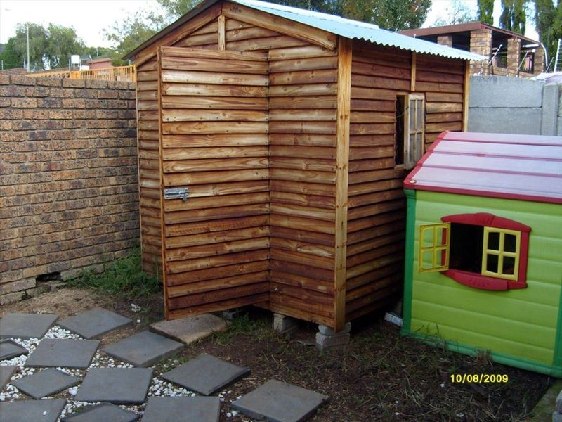 A wooden garden storage shed situated in the backyard, featuring a corrugated metal roof and positioned on a paved area next to a brick wall.