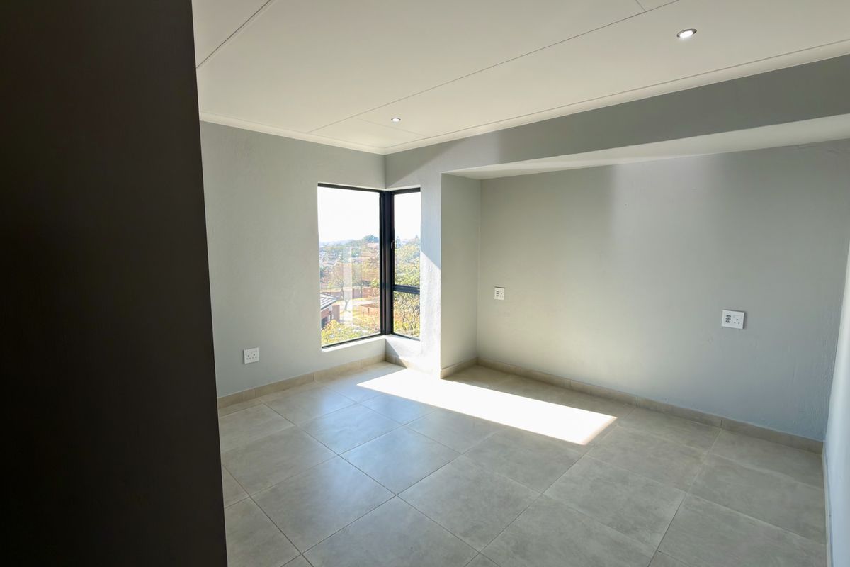 A bright, empty bedroom featuring modern tiled flooring, recessed ceiling lighting, and a large corner window providing natural light and exterior views.