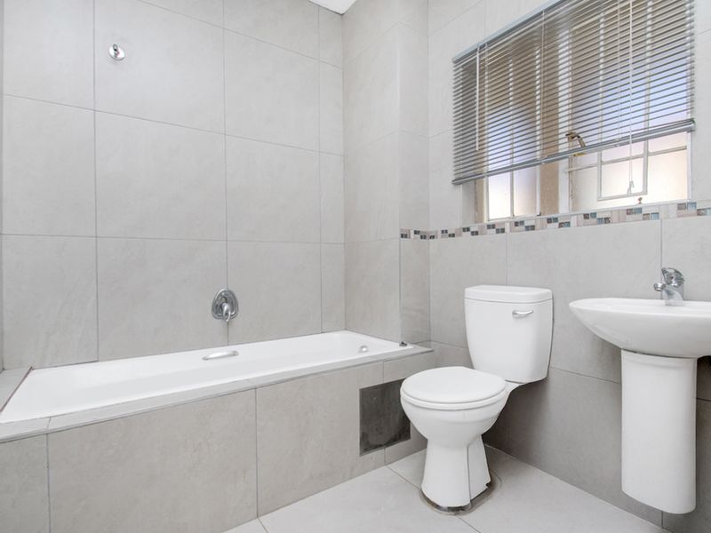 A clean, neutral-toned bathroom featuring a built-in bathtub, pedestal basin, and toilet with modern grey wall tiling and functional window blinds.