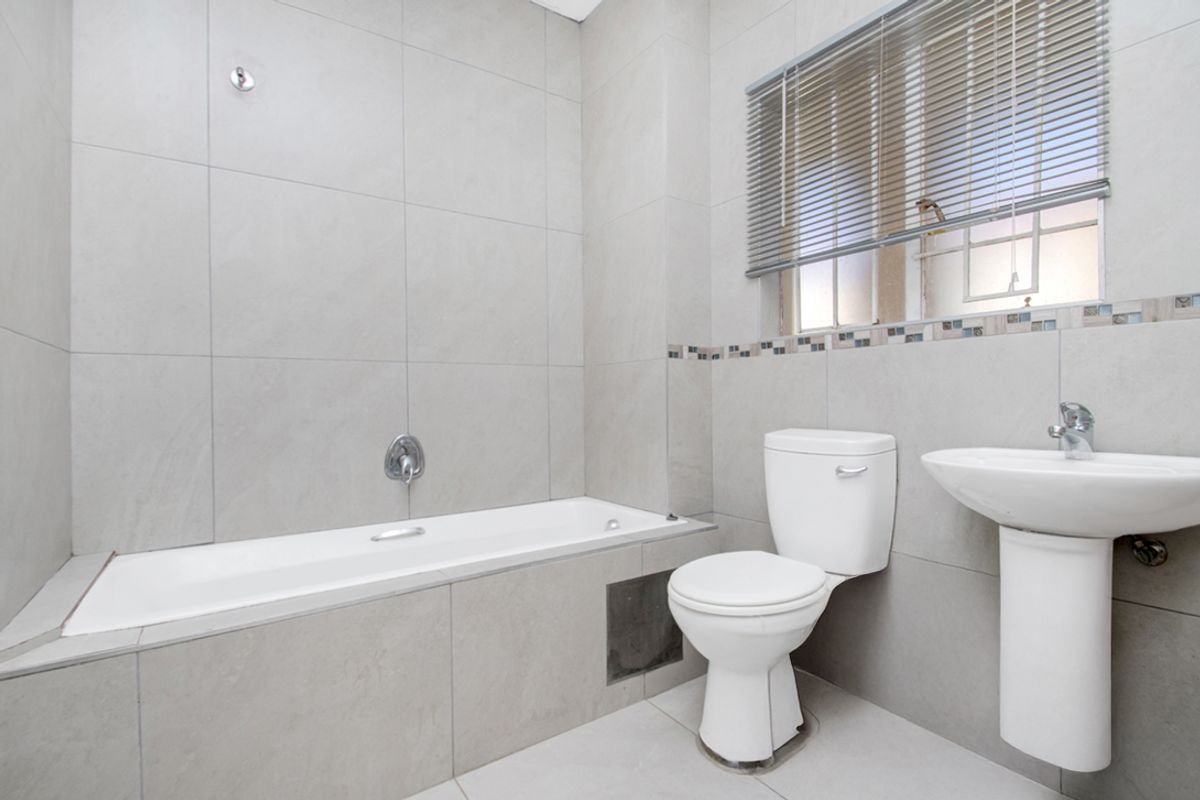 A clean, neutral-toned bathroom featuring a built-in bathtub, pedestal basin, and toilet with modern grey wall tiling and functional window blinds.