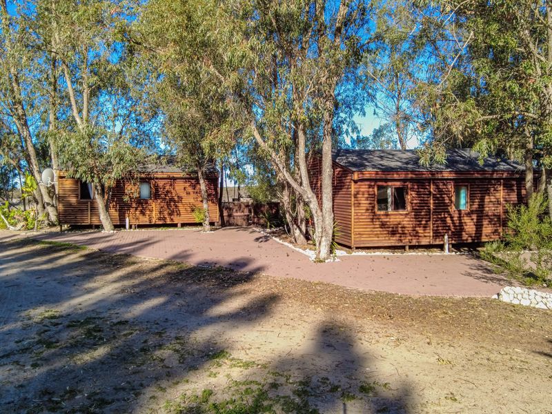 A view of two timber log cabins set within a wooded area, featuring a paved patio space and mature trees.